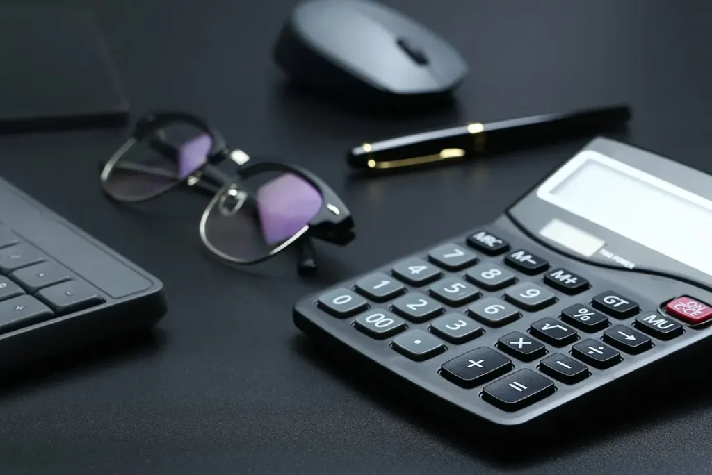 Calculadora, óculos, caneta e mouse, todos pretos ou escuros, sobre uma mesa de tampo preto
