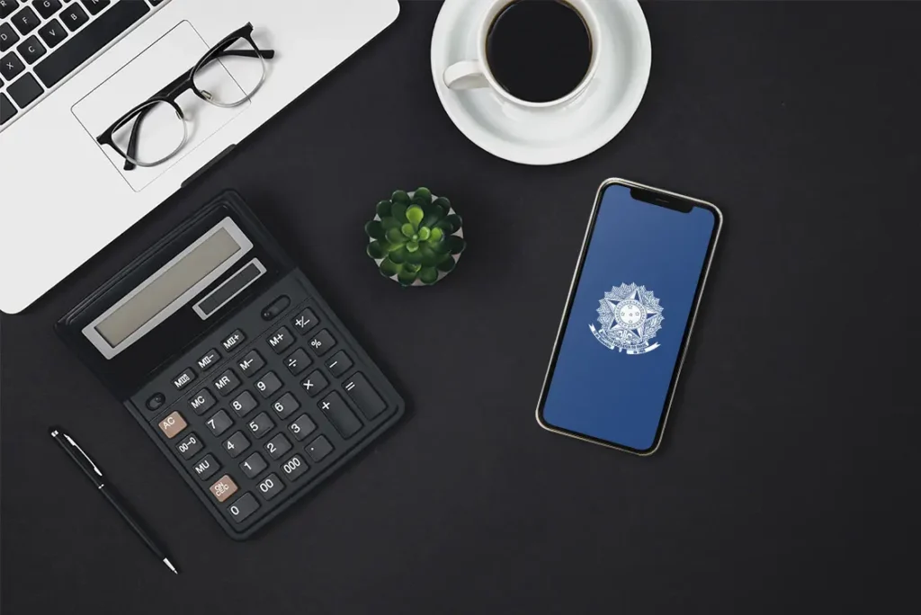 Mesa de trabalho com laptop, calculadora, óculos, café e celular com tela no aplicativo do INSS.