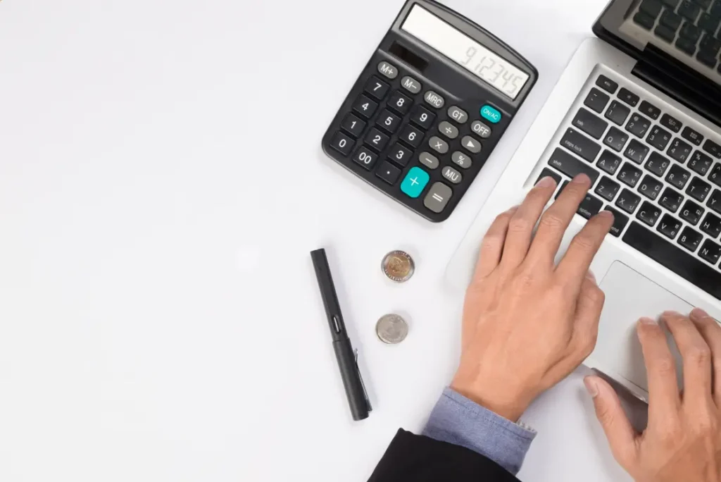 Mãos operando computador sobre uma mesa, com uma calculadora, caneta e moedas ao lado
