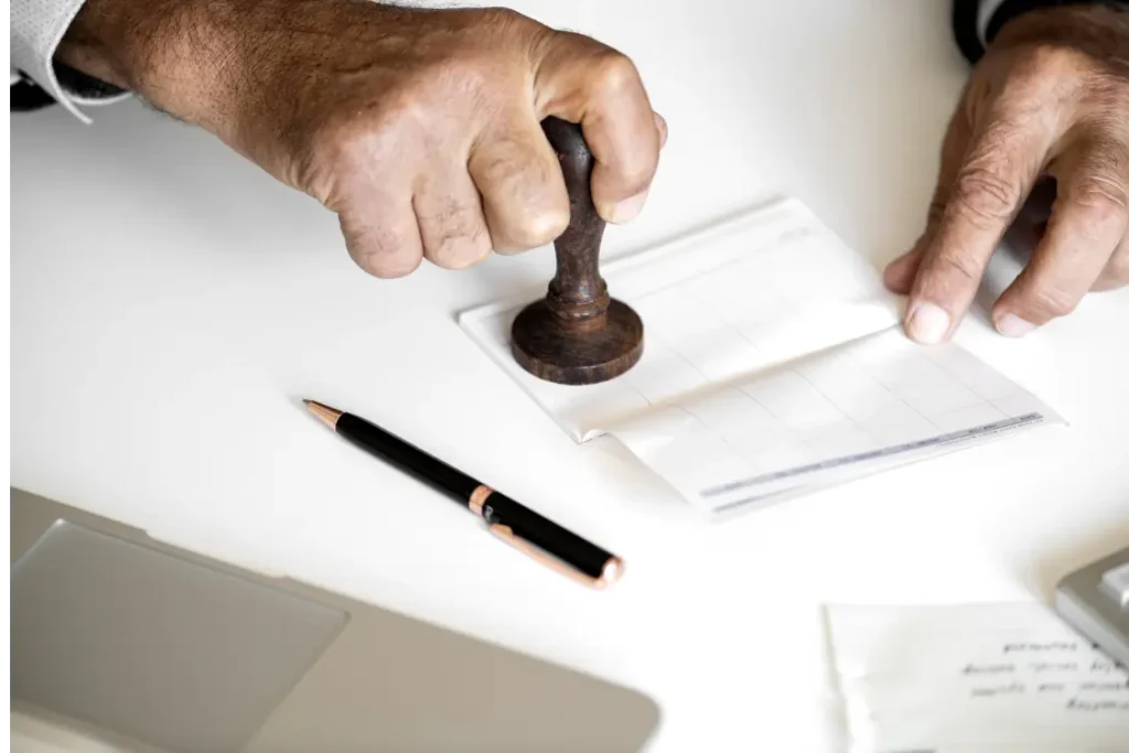 Mão segurando carimbo sobre documento, em uma mesa branca, com uma caneta ao lado