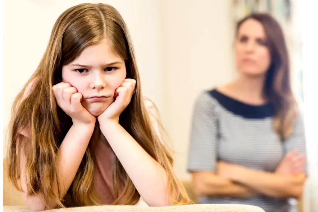 Menina com cara triste e as mãos no rosto. Ao fundo, sua mãe a olha com cara brava e braços cruzados