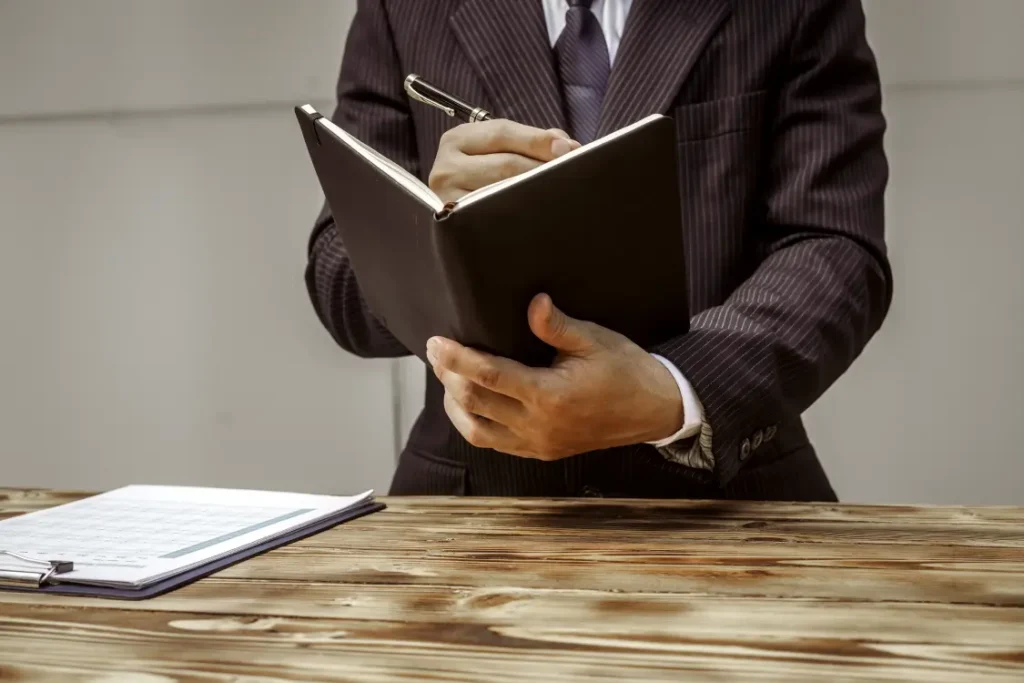 Homem de terno e gravata atrás de uma mesa fazendo anotações em um livro