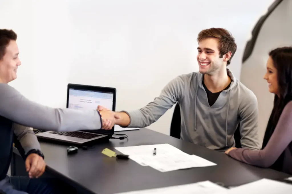 Dois homens, cada um de um lado da mesa, apertam as mãos como símbolo de um acordo fechado. Ao lado de um deles, uma mulher sorridente.