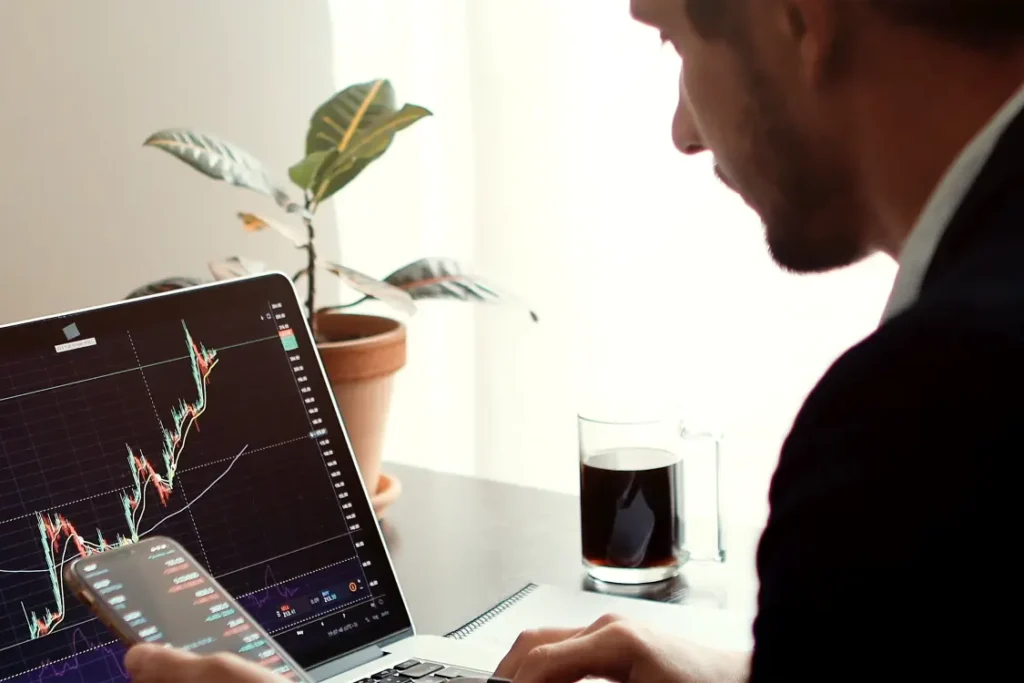 Homem em sua mesa com computador a sua frente e celular na mão, analisando gráficos financeiros.