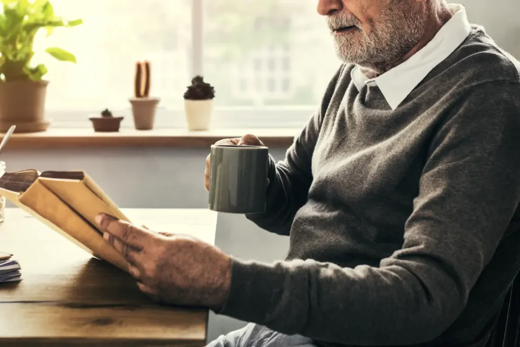 Homem mais velho sentado à mesa, lendo um livro e tomando um café